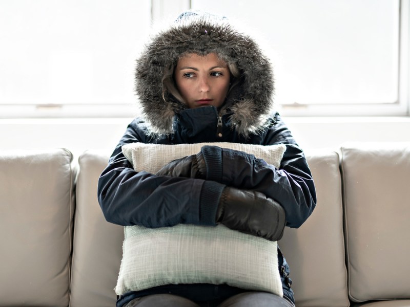 Eine Frau sitzt vollständig mit Jacke angezogen in ihrer Wohnung.