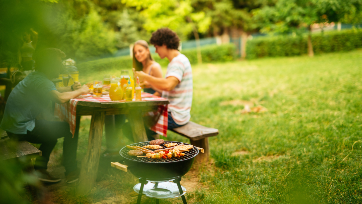 Ein paar Menschen sitzen an einem Tisch und essen. Im Vordergrund sieht man einen Grill mit Grillgut darauf.