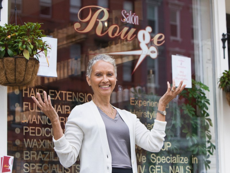 Eine Frau mit kurzen grauen Haaren steht vor einem Friseur-Salon.