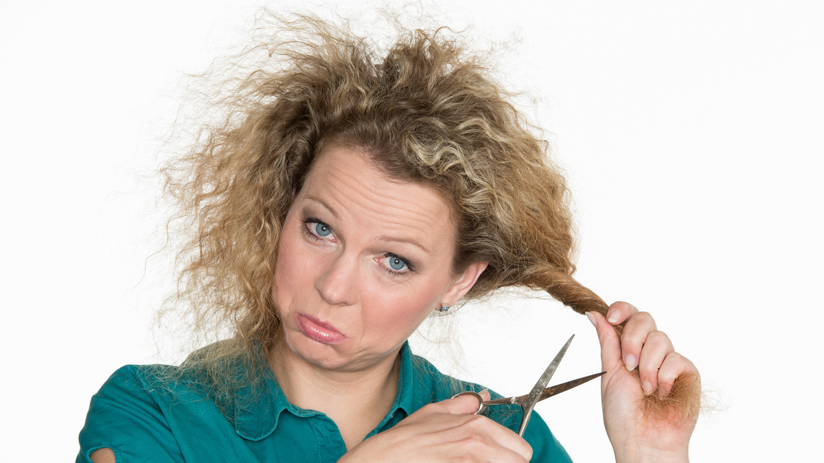 Eine Frau mit Locken vor weißem Hintergrund. Sie hat eine Strähne in der Hand, an die sie eine Schere hält.