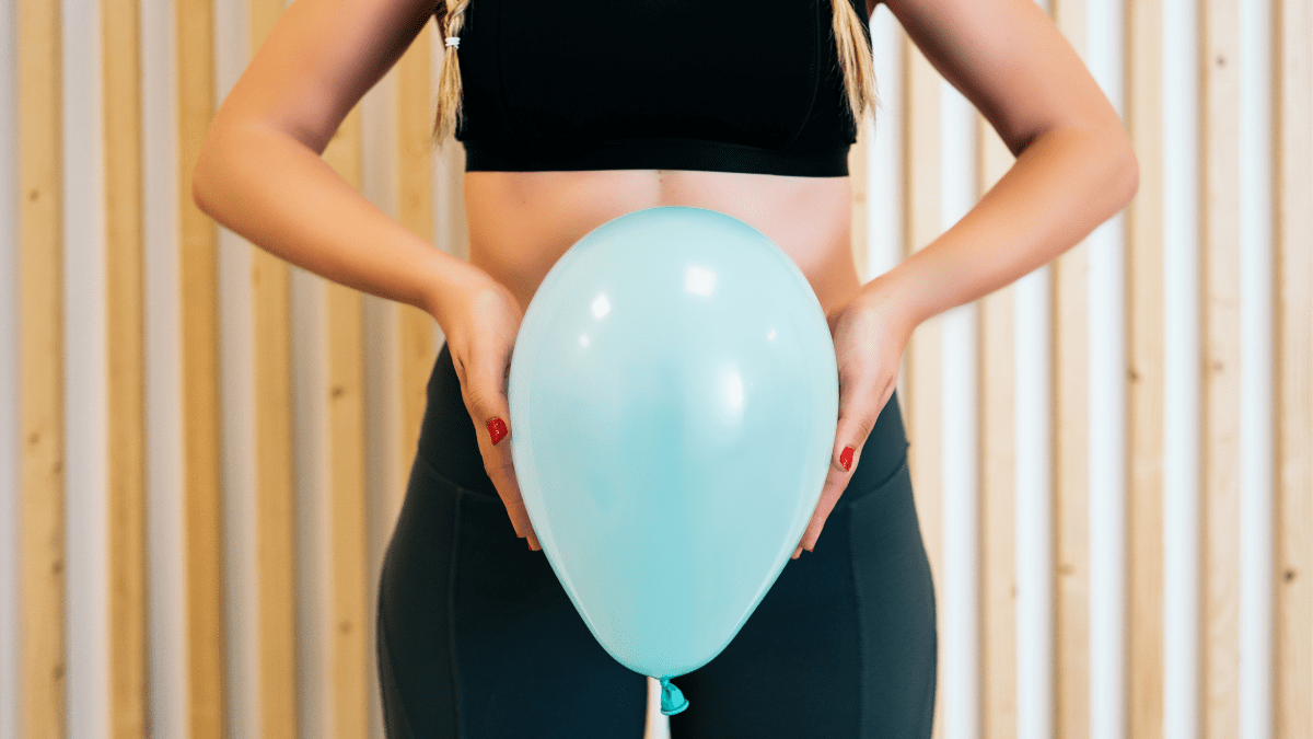 Eine Frau steht mit einem Luftballon vor dem Bauch vor einer Wand.