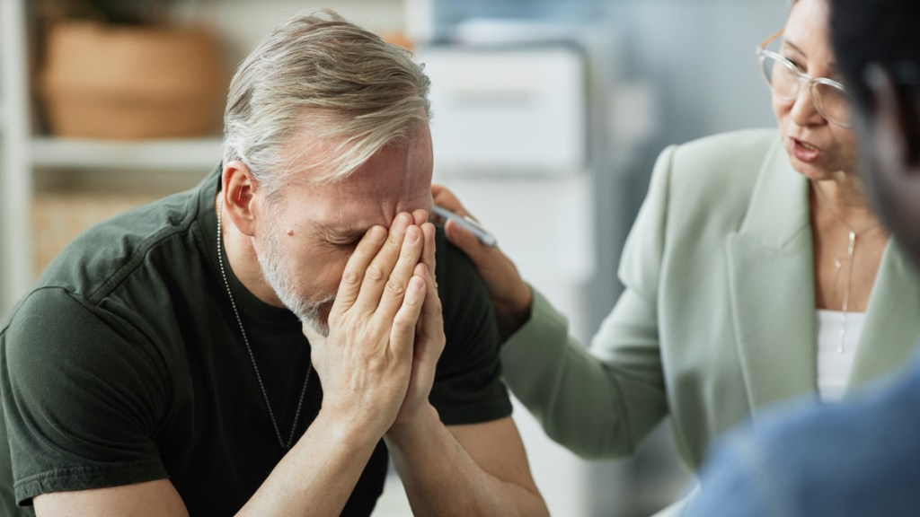 Ein Mann mit kurzen, grauen Haaren hält sich seine Hände vors Gesicht und wird von einer Frau neben sich getröstet.