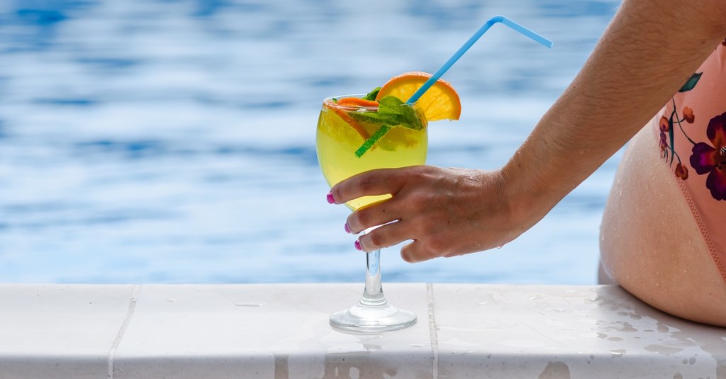 Frau sitzt mit Cocktail am Beckenrand eines Pools.