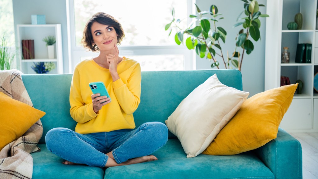 Eine Frau sitzt mit ihrem Handy auf der Couch und überlegt.