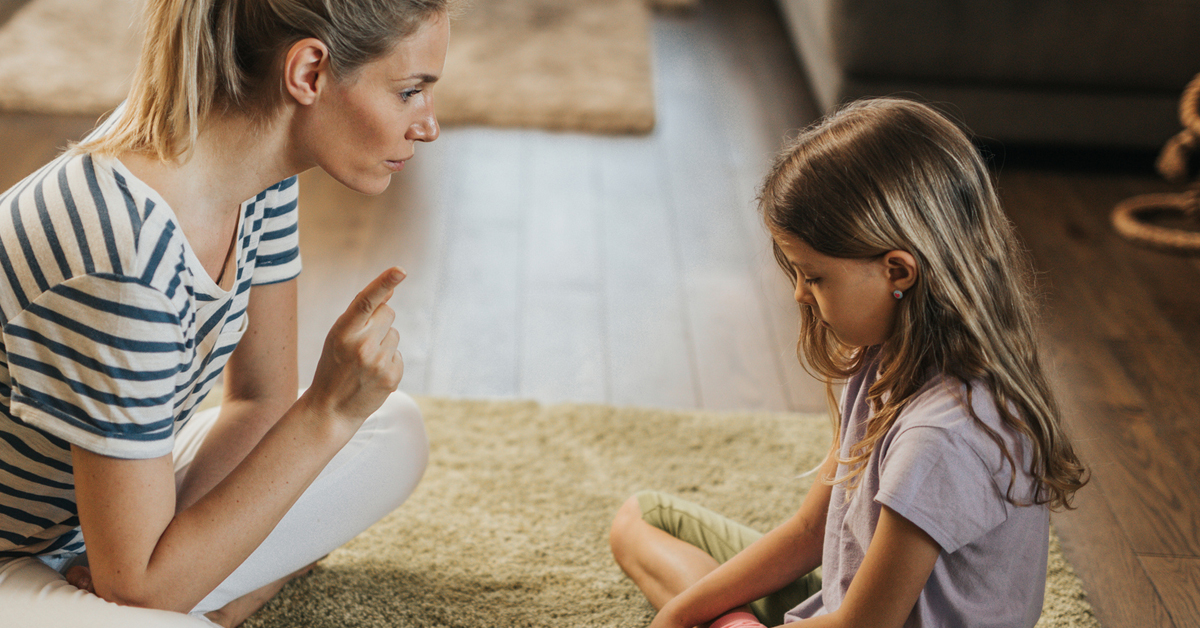 Mutter und Tochter sitzen auf dem Boden. Die Mutter hat den Finger gehoben und tadelt ihre Tochter