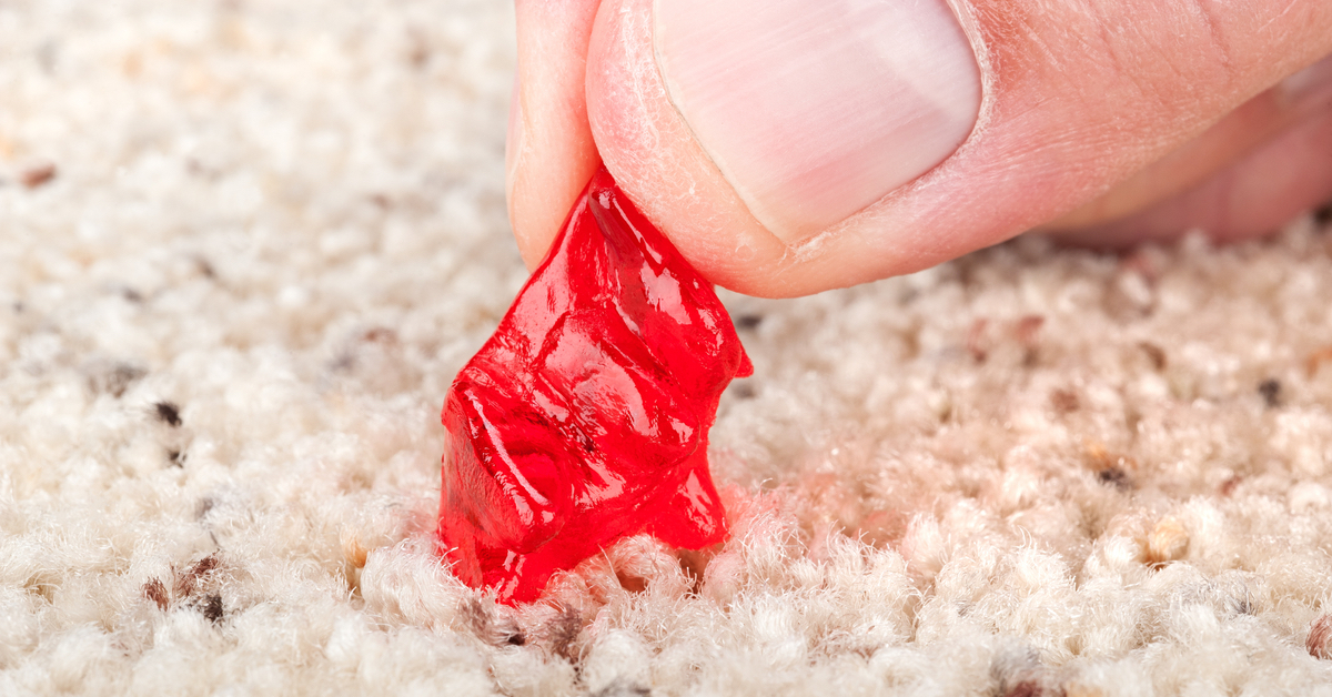 Ein roter Bonbon klebt in hellem Teppich fest.