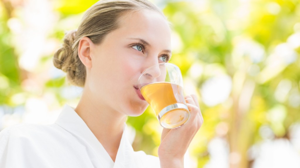 Eine blonde Frau, die unter einem Baum steht, trinkt eine gelbe Flüssigkeit aus einem Glas.