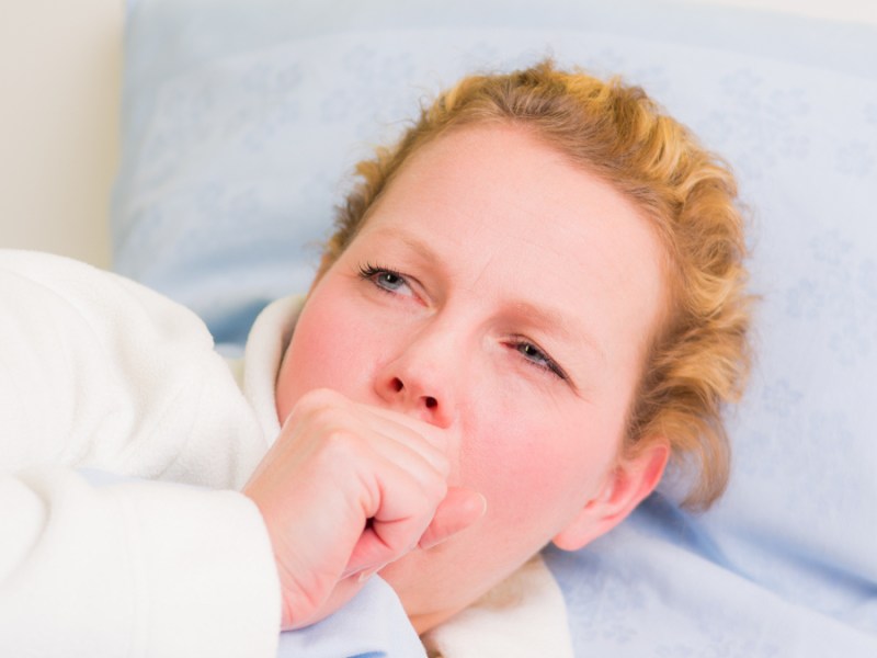 Eine blonde Frau im Bademantel liegt mit Husten im Bett.