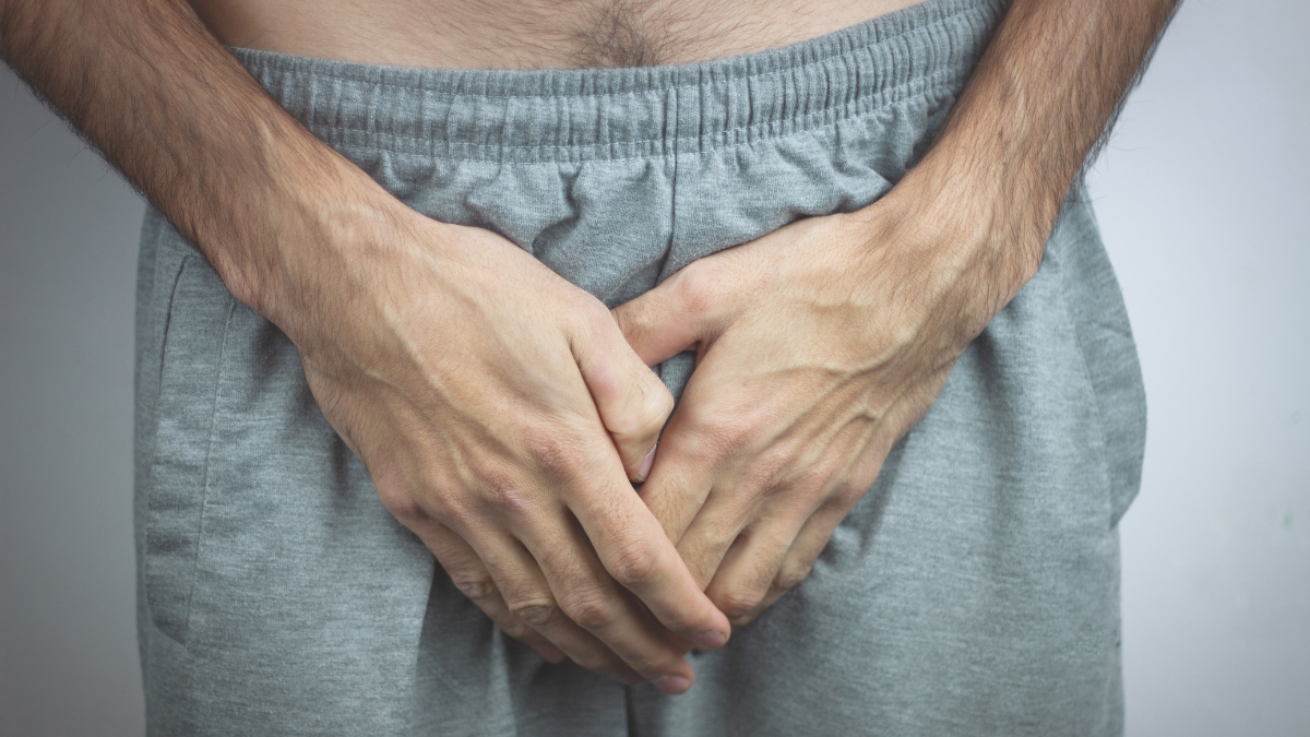 Ein Mann hält auf Höhe seines Geschlechtsteils beide Hände an seine graue Jogginghose.