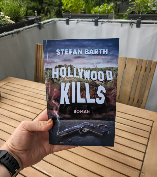 Das Taschenbuch des Romans „Hollywood Kills“ wird in die Kamera gehalten.