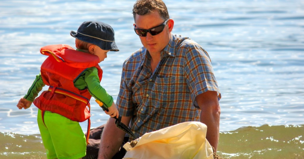 Ein Vater sitzt mit seinem Sohn am Strand, der Sohn trägt Badebekleidung, einen Hut und eine Schwimmweste