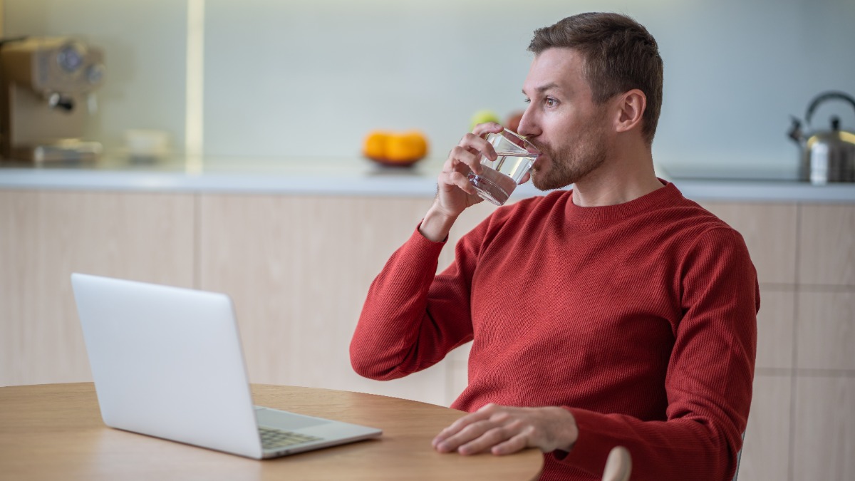 Ein Mann sitzt vor seinem Laptop und trinkt ein Glas Wasser