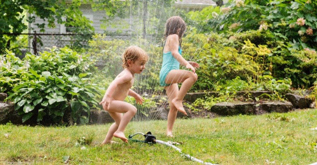 Nackiger Junge und seine große Schwester, die einen Badeanzug trägt, springen über einen Rasensprenger.