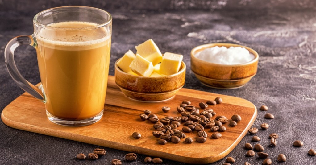 Ein Glas mit einem Bulletproof Coffee, darum verteilt liegen Kaffeebohnen, Butterstückchen und Kokosöl.