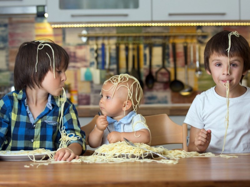 drei Kinder sitzen am Tisch und haben Spaghetti auf dem Kopf