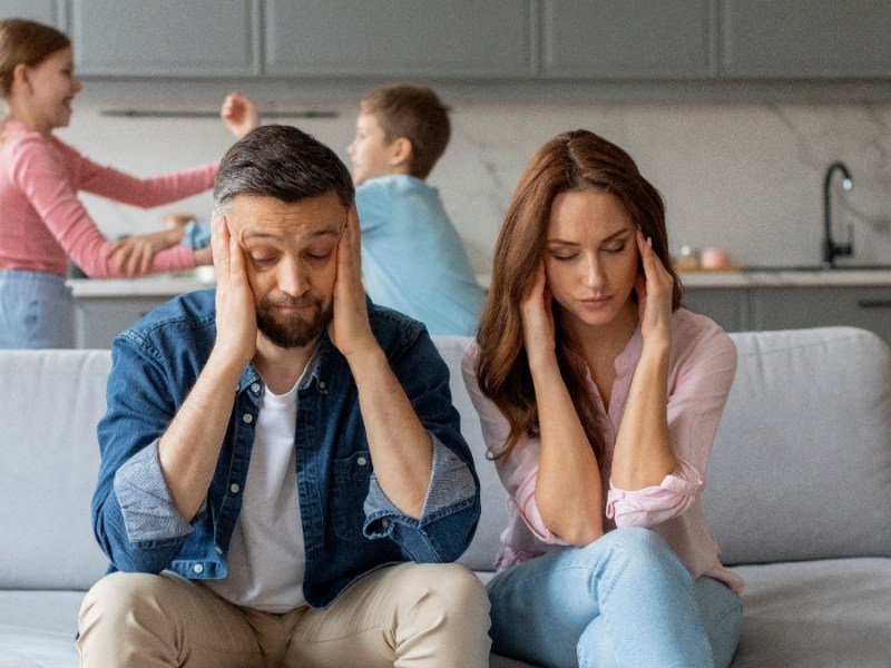 Eltern sitzen genervt auf der Couch, während sich die Kinder im Hintergrund streiten