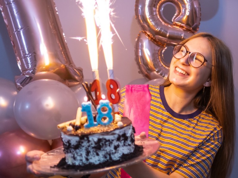 ein Mädchen hält eine Torte in der Hand, im Hintergrund sind zwei Luftballons zu sehen, die eine 18 zeigen