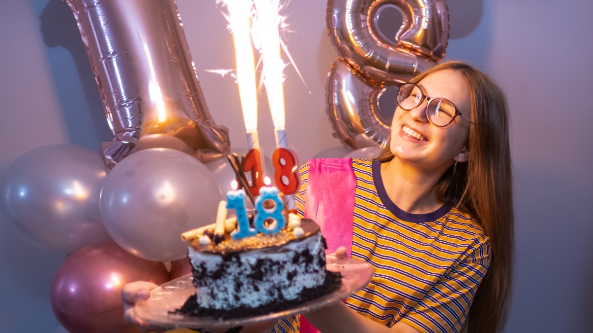 ein Mädchen hält eine Torte in der Hand, im Hintergrund sind zwei Luftballons zu sehen, die eine 18 zeigen