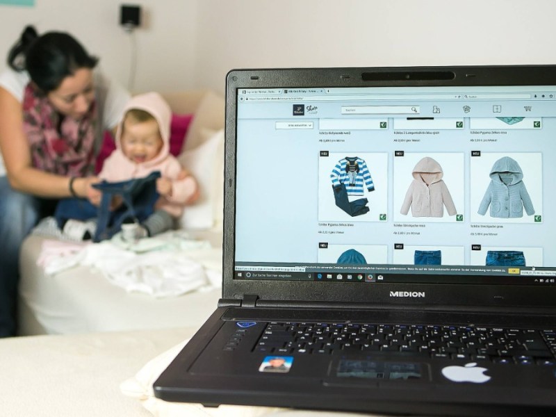 Ein Laptop mit einer Seite für Kindermode ist zu sehen, dahinter sitzt eine Mutter mit ihrem Baby auf dem Sofa