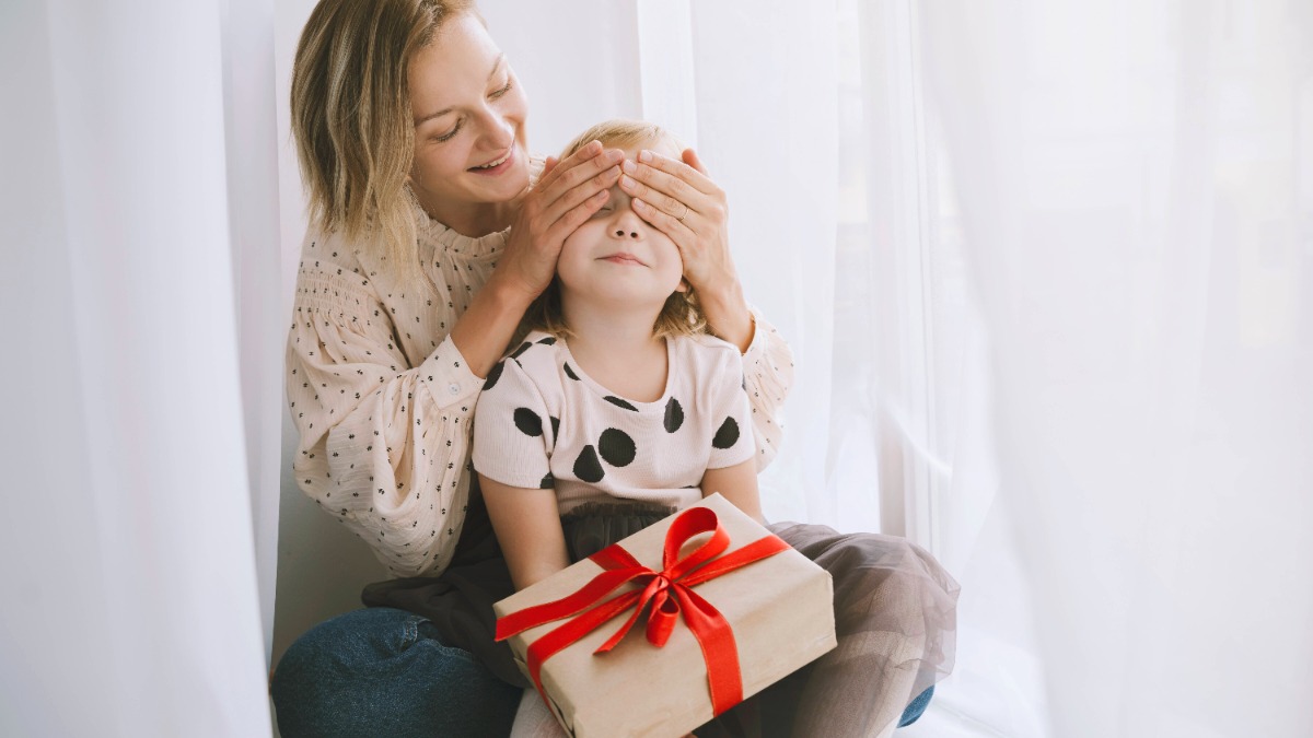 Alleinerziehend an Weihnachten: Mutter verdeckt die Augen ihrer Tochter, auf deren Schoß ein Geschenk liegt