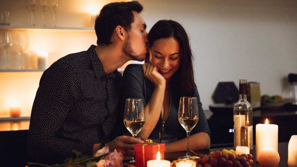 Ein heterosexuelles Pärchen sitzt bei Kerzenschein auf der Couch. Er küsst ihre Schläfe und sie lacht.