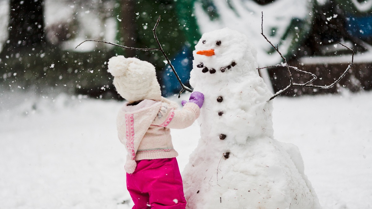 Schneemann bauen: ein kleines Mädchen steht vor einem Schneemann